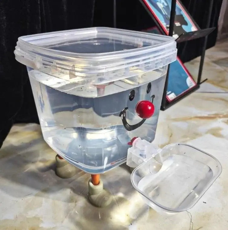 Clear plastic automatic gravity pet water dispenser with a smiling face, red nose, and pipe fitting legs