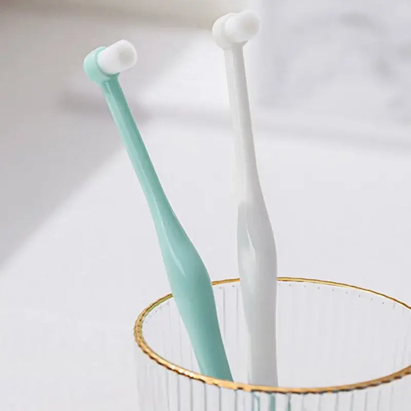 Two mint green and white mini dog toothbrushes with soft rounded bristles in a clear glass cup with gold rim for oral care
